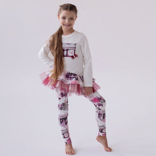 Young girl wearing a white long-sleeve top  with a maroon tulle hem and maroon patterned leggings on a light gray background