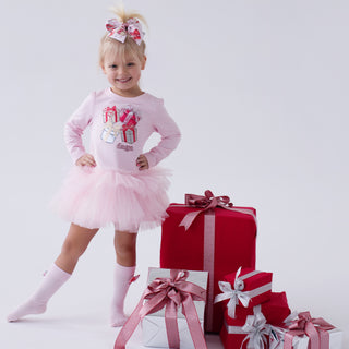 Child in pink Daga Magical Surprises  outfit with a bow, standing next to wrapped gifts on a white background