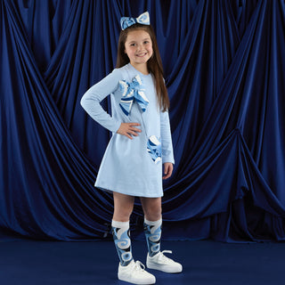 Young girl wearing a light blue dress with a pattern, standing against a dark blue curtain.